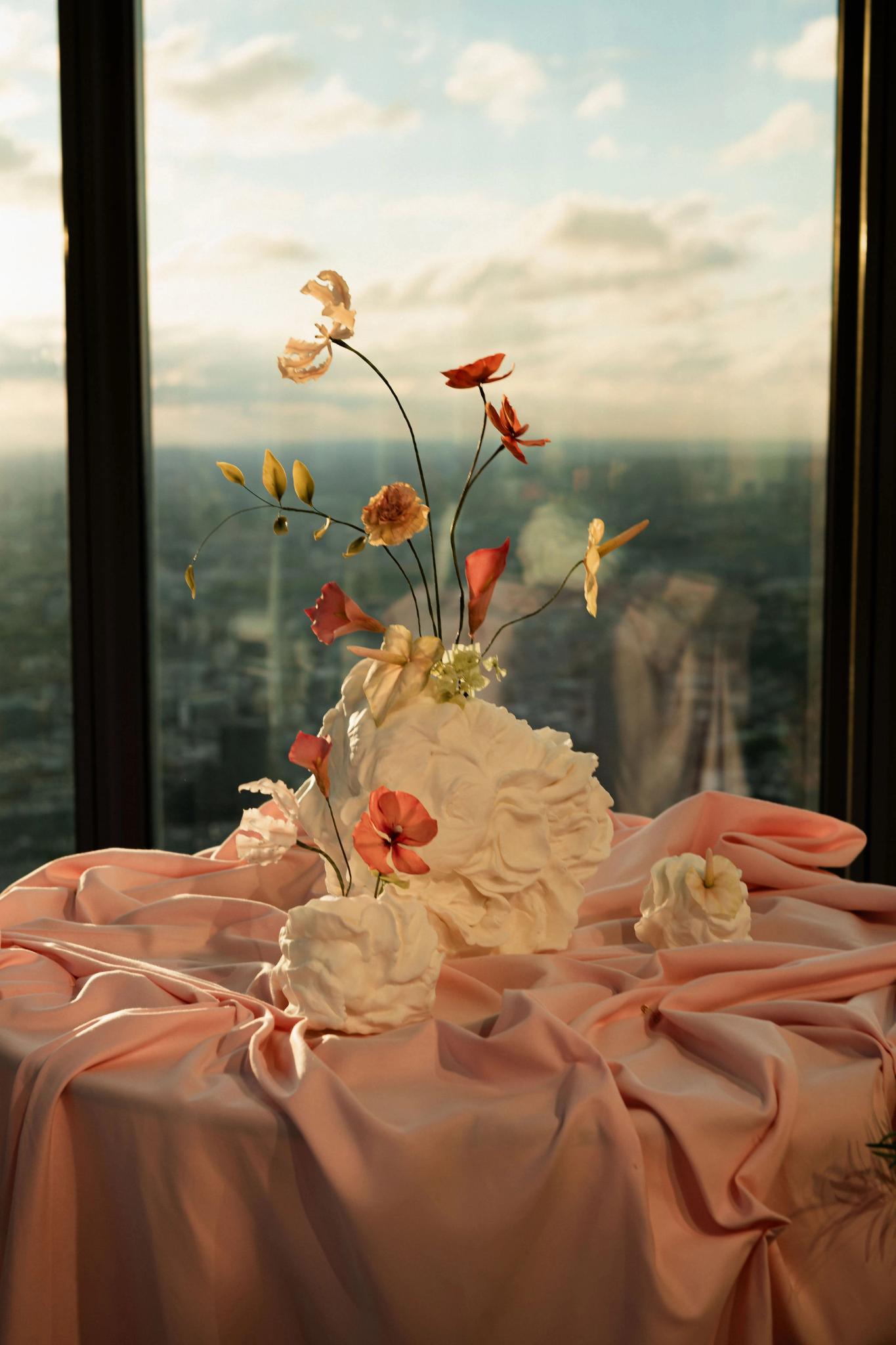 A floral arrangement featuring sculpted white flowers and delicate pink and yellow blossoms displayed on soft pink fabric, with city buildings and a sunset sky visible through large windows in the background.