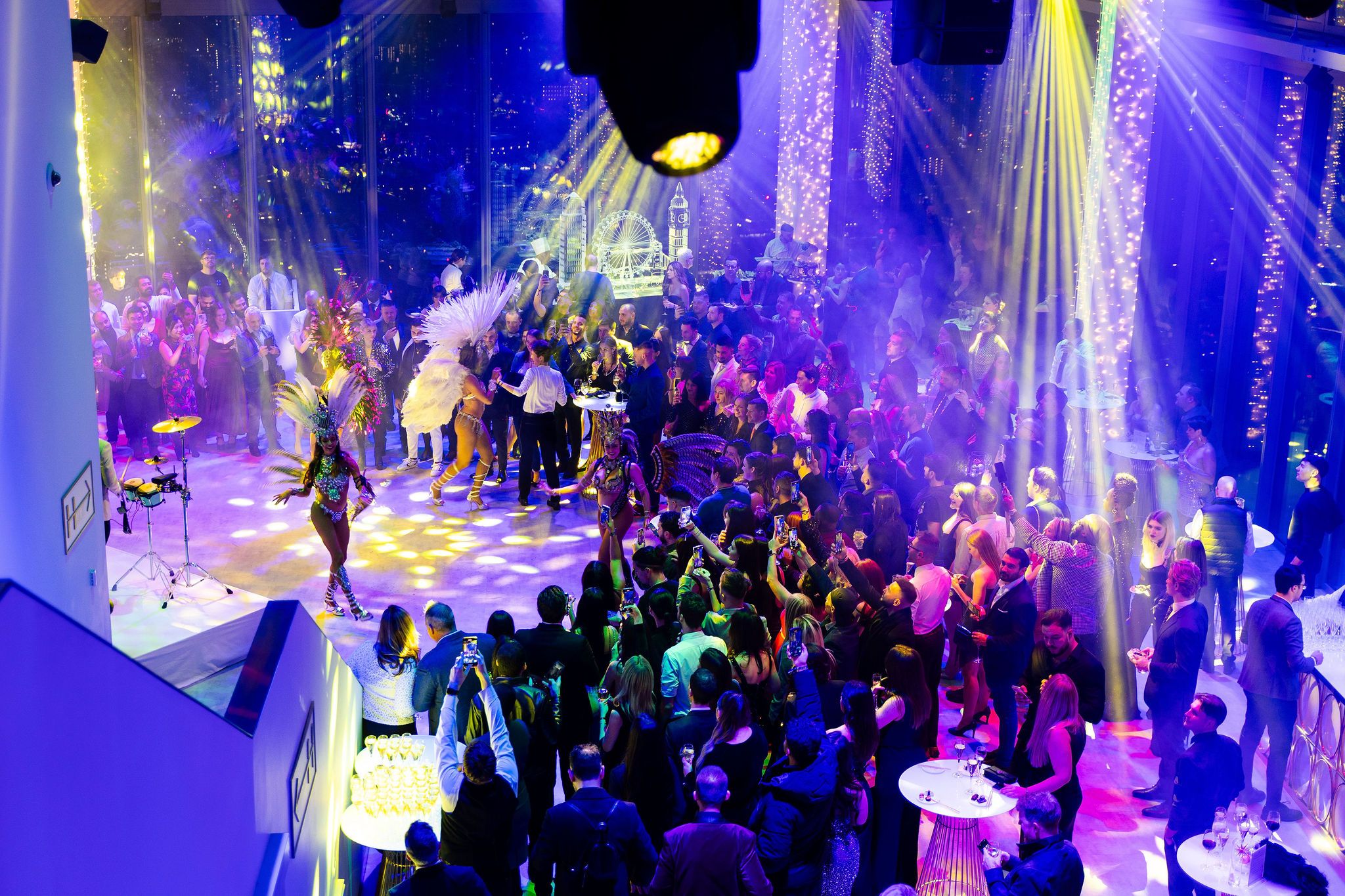 Distant view of dense pink and red fireworks across a cityscape at night, framed by dark vertical window bars and silhouettes of onlookers.