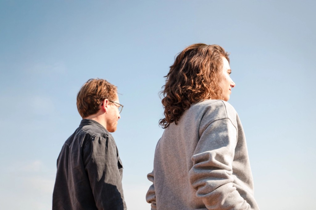 Eine weiblich und eine männlich gelesene Person gehen im Raum mit großen Fenster und Holzbalken am Boden.
