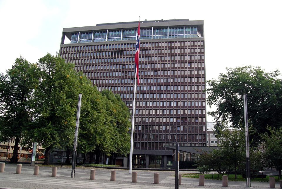 Flagpole with Norwegian flag in place with trees in front of office building.