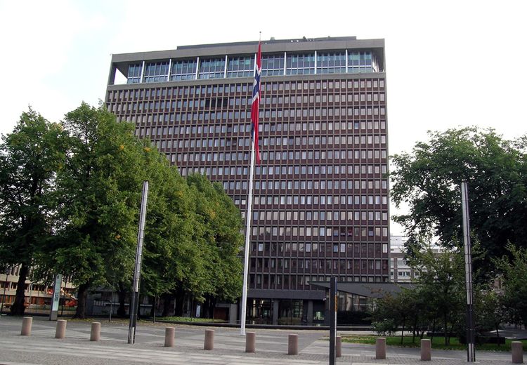 Flaggstang med norsk flagg på plass med trær fremfor kontorbygning.  