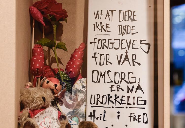 Exhibition display with different objects, textile flowers, teddy bears, a brickstone, a skateboard with the text: "Know that you did not die in vain. Our care is unwavering. Rest in peace".