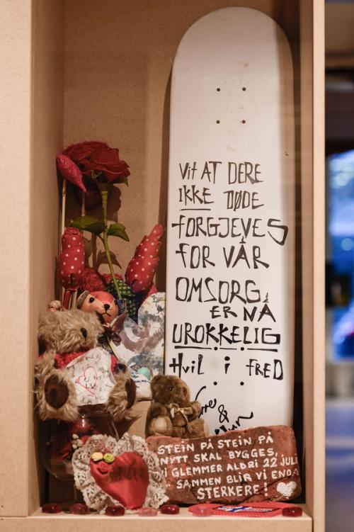 Exhibition display with different objects, textile flowers, teddy bears, a brickstone, a skateboard with the text: "Know that you did not die in vain. Our care is unwavering. Rest in peace".