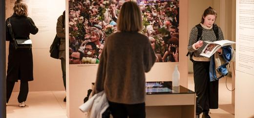 Three female visitors seen from behind read some tekst boards in an exhibition. Another female visitor reads a book next to one of the boards in the exhibition. In the middle there is a board with a large format picture of people holding roses.