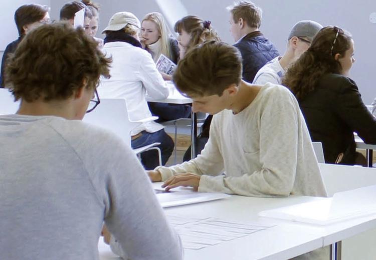 Et titalls unge mennesker sitter på hvite stoler ved hvite bord og skriver og snakker sammen. Hvite vegger i bakgrunnen.