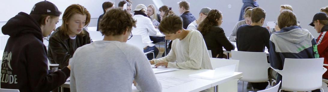 Et titalls unge mennesker sitter på hvite stoler ved hvite bord og skriver og snakker sammen. Hvite vegger i bakgrunnen.