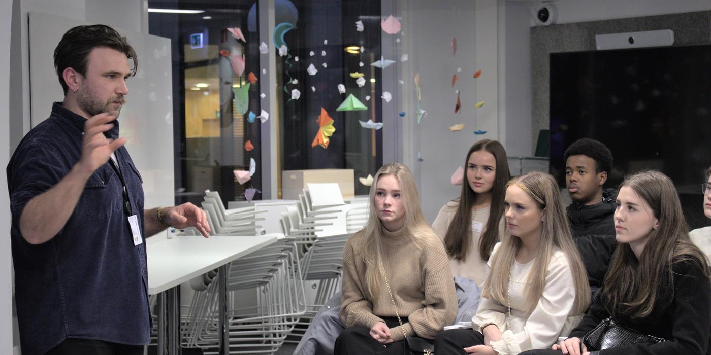 A man stands and speaks to a group of students sitting in a classroom. At the back is a white table and several white chairs, in front of a large window with origami figures hanging from the ceiling.