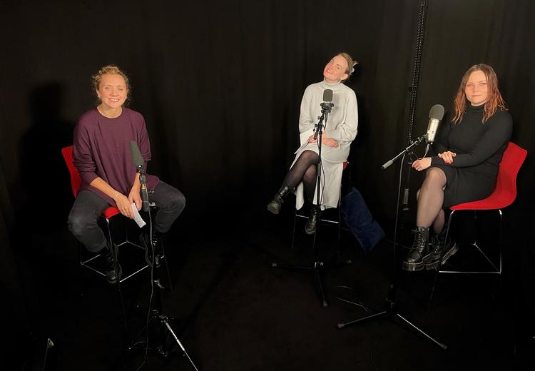 Tre smilende pene kvinner sitter på røde stoler med mikrofoner på stativ foran seg.  Sort bakgrunn. Foto