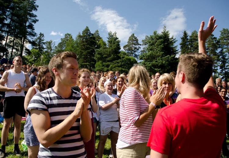 Picture of a boy in a red t-shirt raising his hand to thank the applause from the young people around him. They are standing in a clearing and it is nice summer weather.