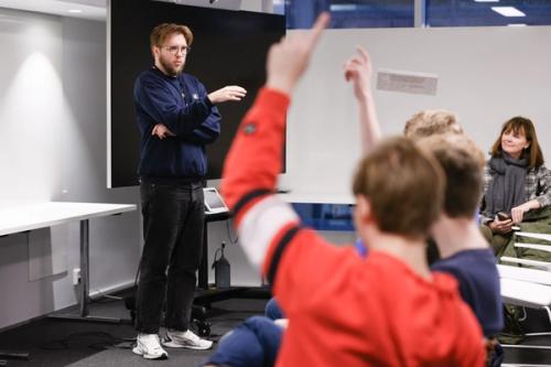 A man stands in front of seated students seen from behind. Two students raise their hands.