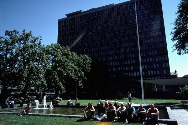 Large Highrise building, in the foreground a group of people sitting in front of a fountain. Trees to the left in the photo.  