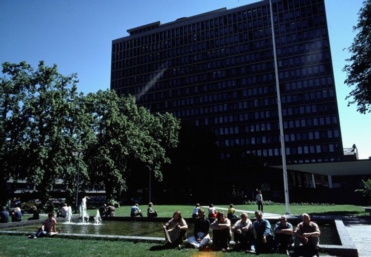 Large Highrise building, in the foreground a group of people sitting in front of a fountain. Trees to the left in the photo.  