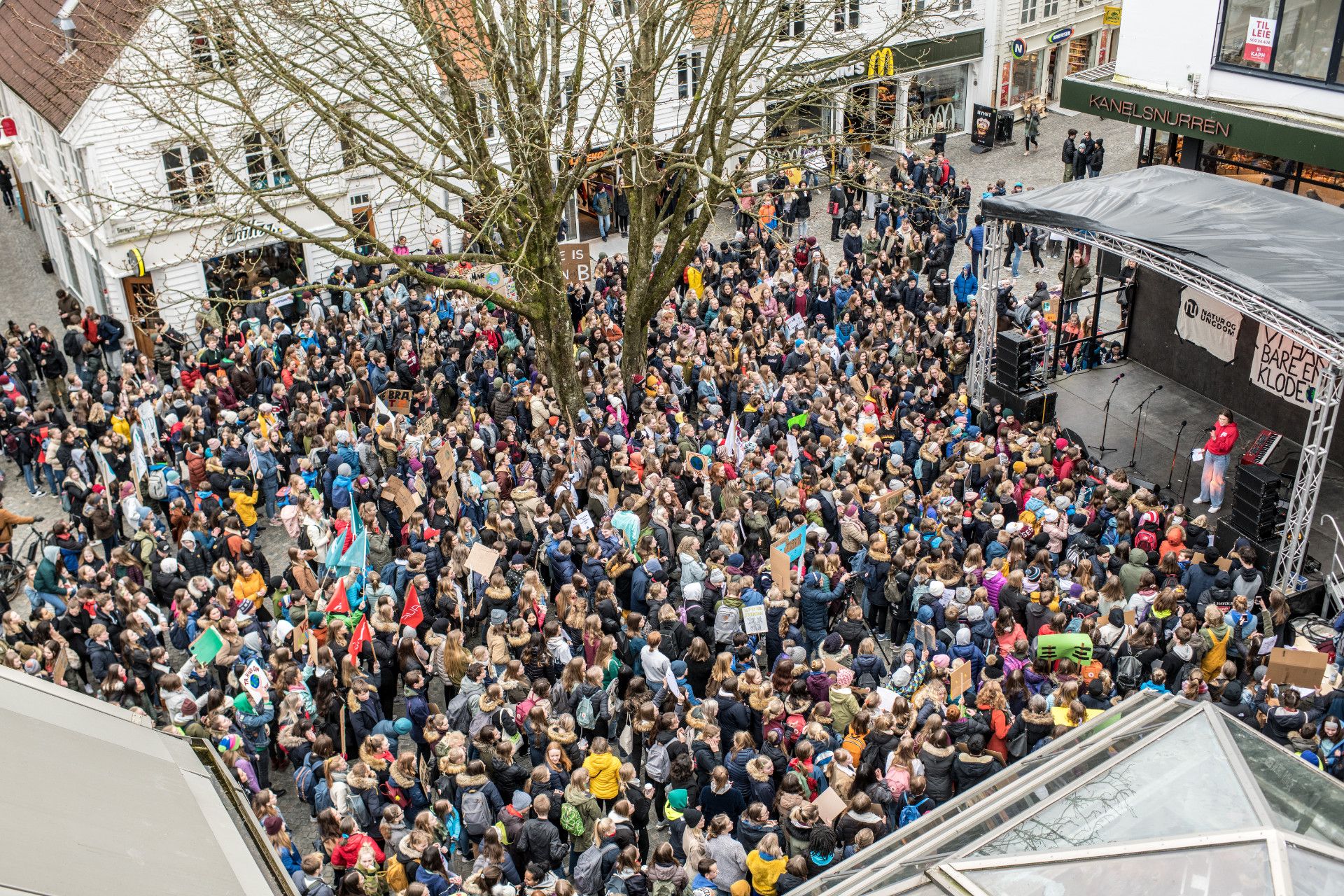 Overview of dense crowd in street. An outdoor scene with a person with a microphone. On the stage hangs a banner with an inscription "VI HAR BARE EN KLODE" and "NATUR OG UNGDOM". 