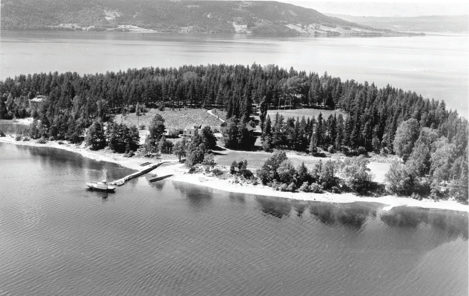 Black-and-white aerial photo of a tree-dense island with wooden house, dock and boat. Quiet water. Landscape with trees in the background.
