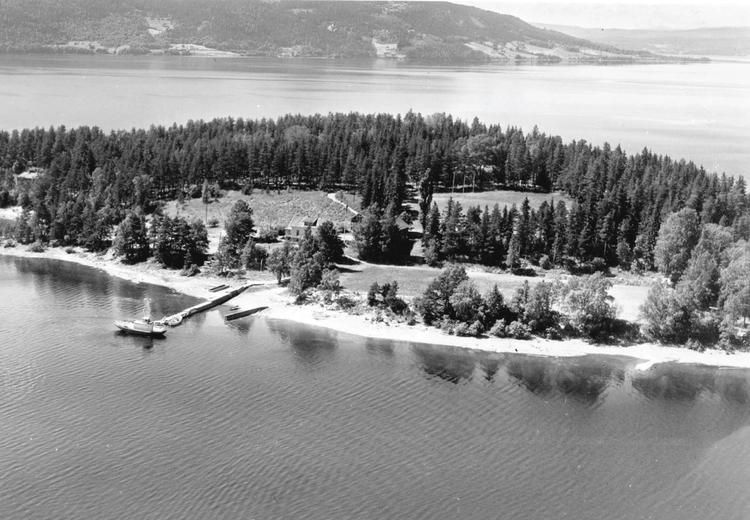 Flyfoto av svart-hvitt av tretett øy med trehus, kai og båt. Stille vann. I bakgrunnen landskap med trær.