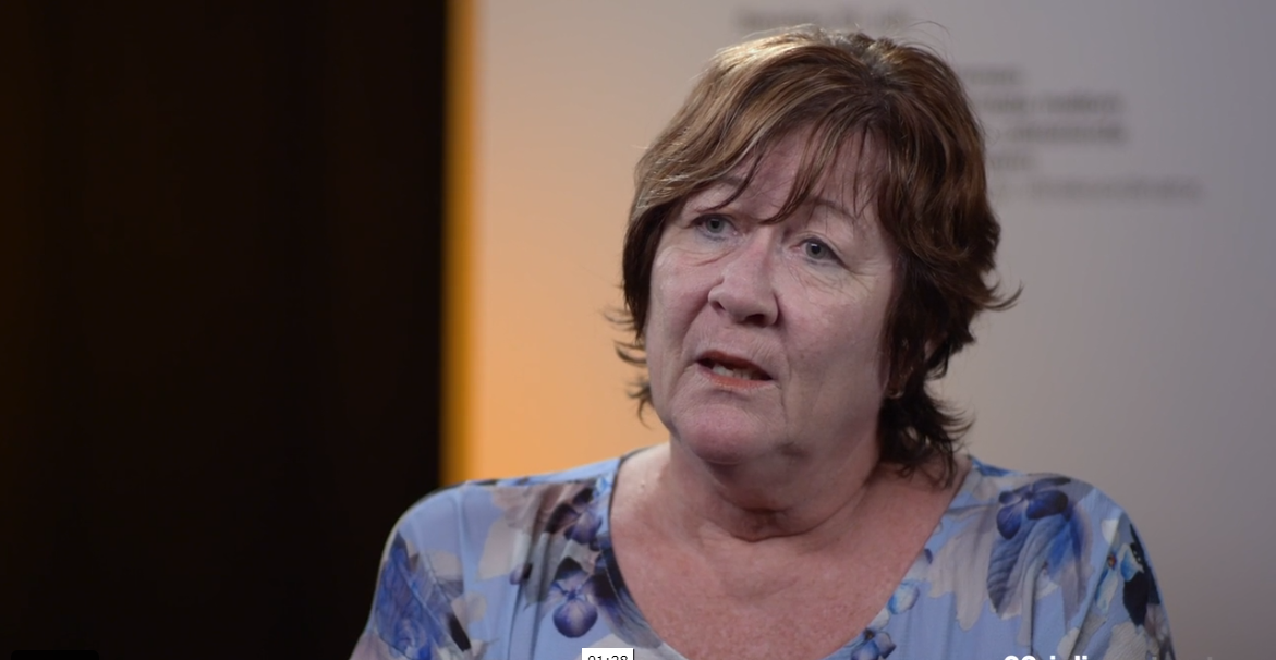 Woman with short brown hair talking. Blurred background.