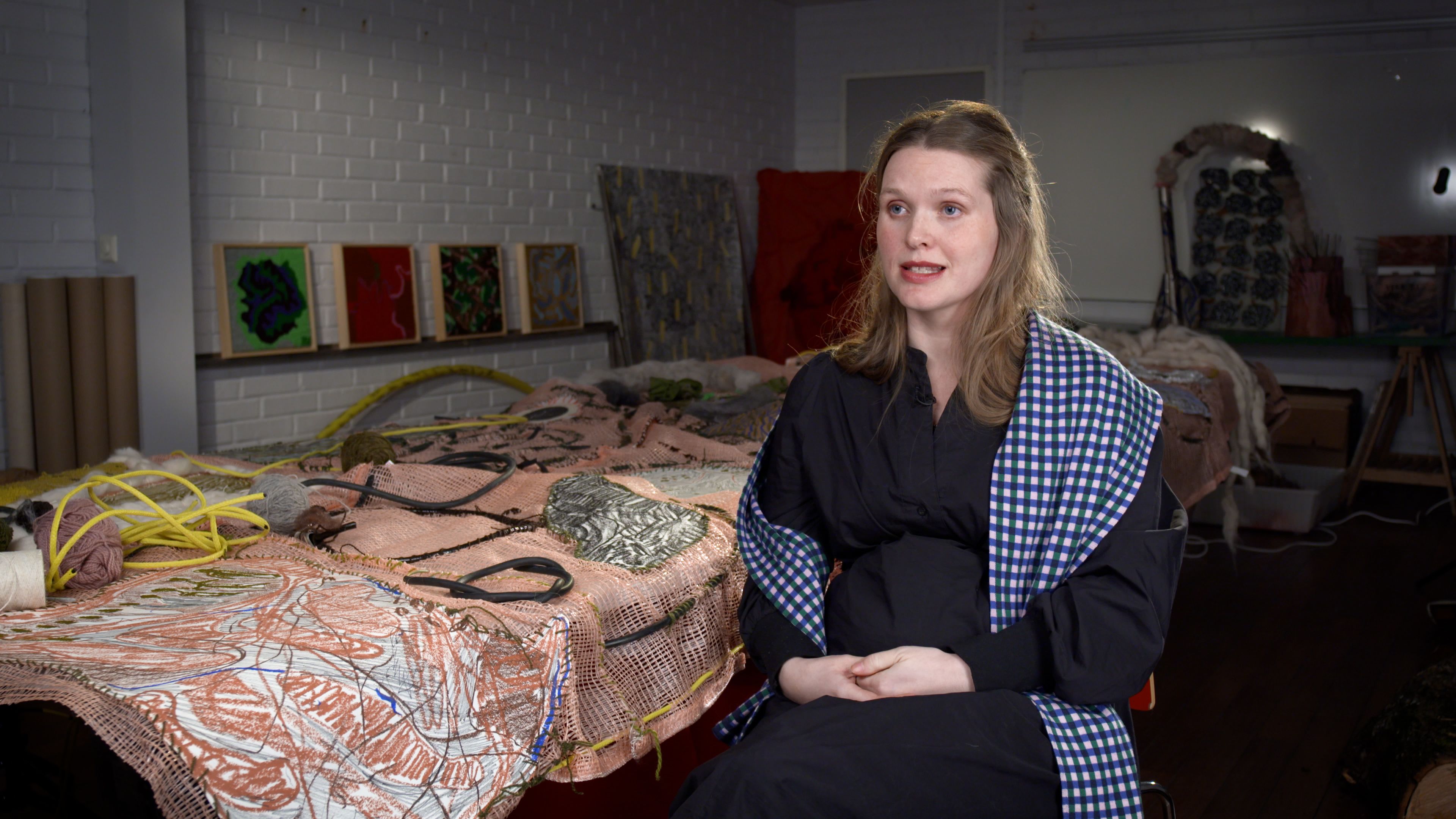 Woman in a black dress, sitting on a chair besides a large piece of artwork on a table.