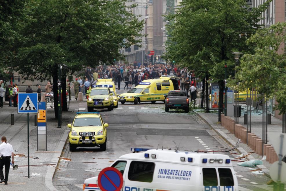 I forgrunnen står en politibil. Lenger bak fire ambulanser. Bakerst i bildet en stor folkemengde. Foto