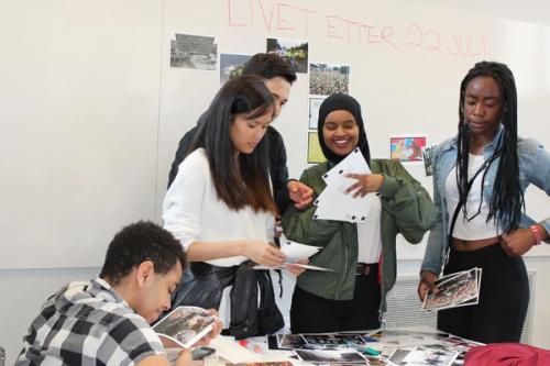 Five people holding pictures. Unclear motives on the pictures. Several pictures lying on the table in front of people. Board in the background with a photo hung on the board.