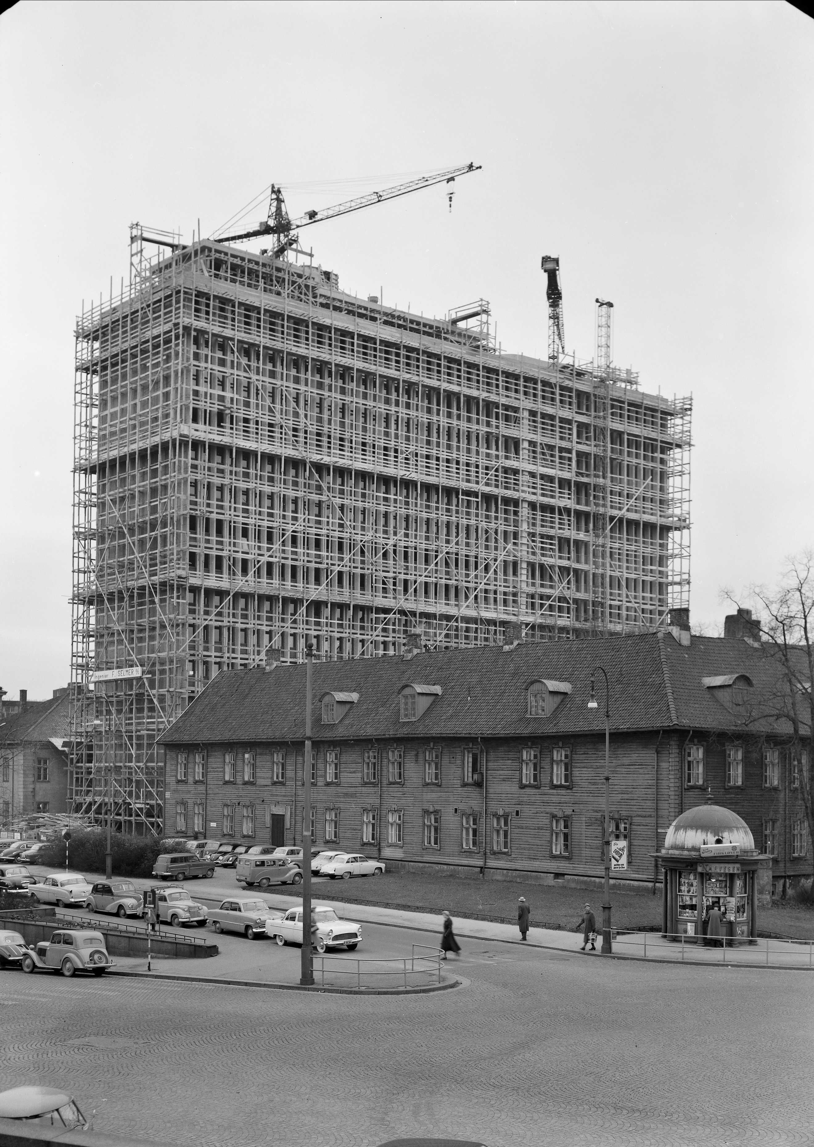 Black and white image of building under construction. An older low building. Cars. Menneker walks by.