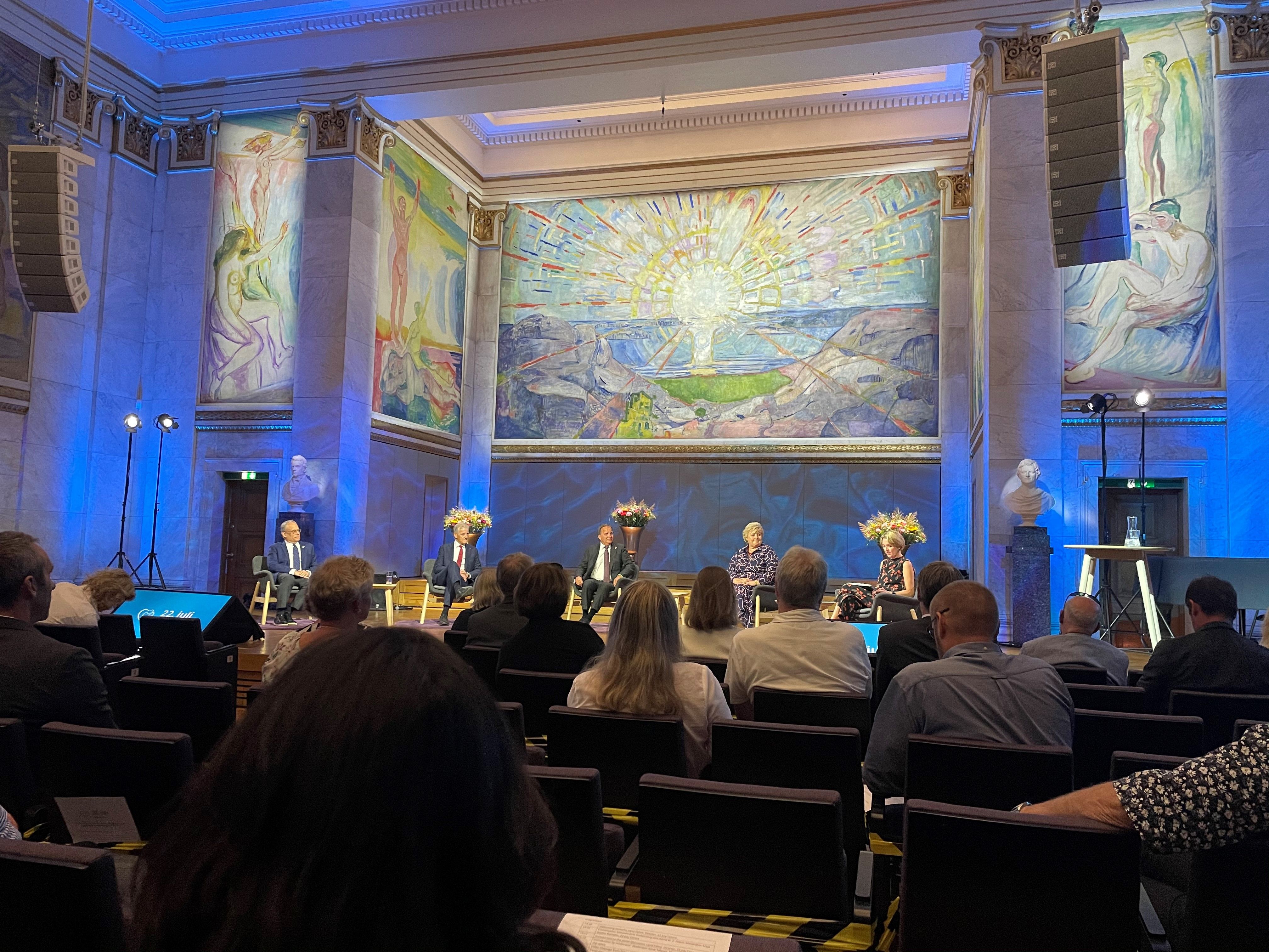 Five people sit on a stage. Blue background with large-format expressionist paintings. The audience sits in front of the stage.