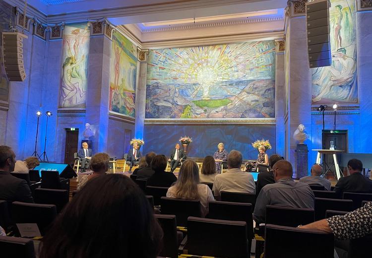 Fem mennesker sitter i en panel. Blå bakgrunn med storformat ekspresjonistiske malerier. Publikum sitter foran scenen.