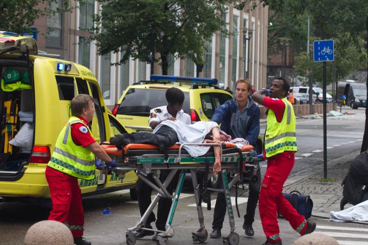 Fire mennesker triller en båre med en skadd person. En ambulanse og en legebil. Bygning i bakgrunnen