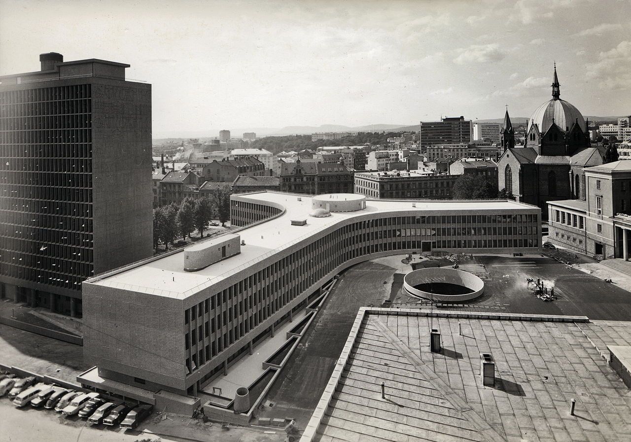 Black and white photo of an urban landscape. In the middle, a modernist building that has a Y format. Next to the building is a high-rise block. Several cars are parked in front of the two buildings.
