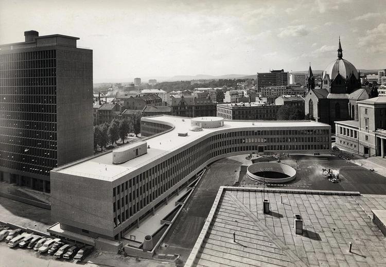 Bilde i sort/hvitt av et urbant landskap. I midten er en modernist bygning som har en Y-format. Ved siden av  bygningen  er det en høyblokk. Flere biler er parkert foran de to bygningene.