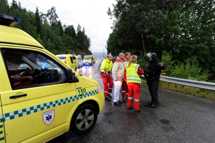 Yellow ambulance at the front of the picture. Five people. Three ambulances further back in the picture. Wet asphalt. Road. Trees