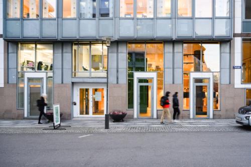 Building facade with windows and large glass surfaces. Asphalt road ahead. Three walking people seen out of focus.