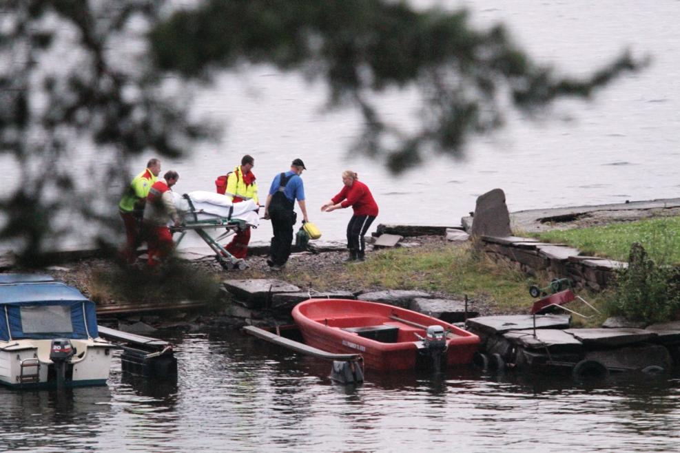 Blurry tree in the foreground. A white boat and a red boat lying at dock. Five people transfers a stretcher with an injured person. Water in front and behind the dock