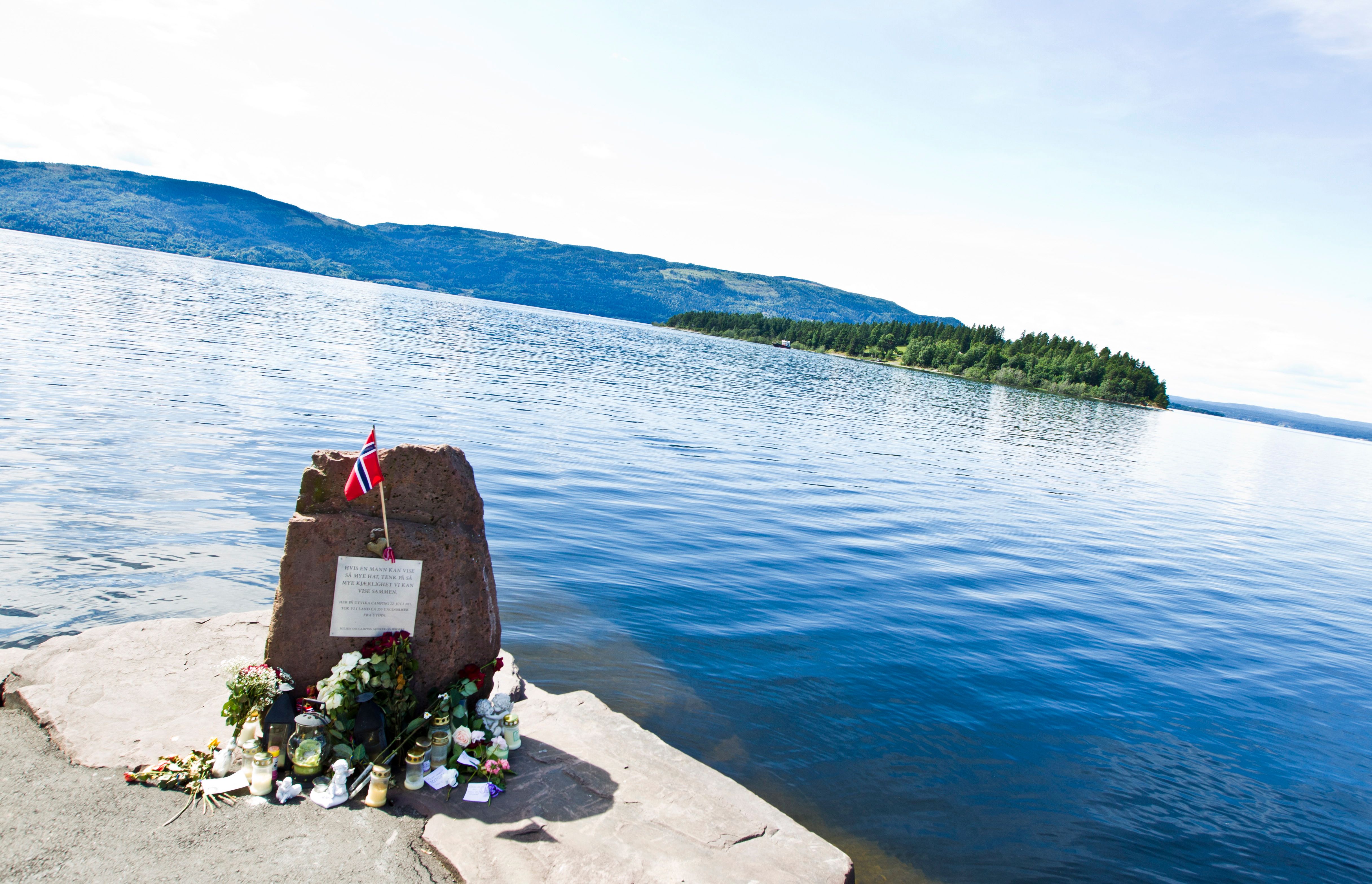 Memorial stone with inscription. Lights, flowers and flags around the stone. A water with a green island and land on the other side of the water. Photo