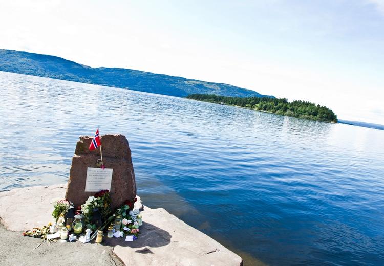 Memorial stone with inscription. Lights, flowers and flags around the stone. A water with a green island and land on the other side of the water. Photo