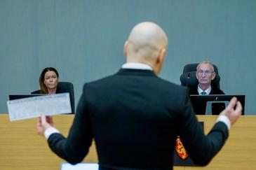 Snauklipt person med dress, terroristen, står med hendene utslått. Man ser ryggen til personen. I bakgrunnen sitter to personer bak et bord og ser på mannen.