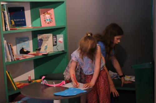 A child draws on a white sheet of paper with a red pencil on a round small table. Behind the child another child leans and sees something. On the left, a green bookcase with children's books.