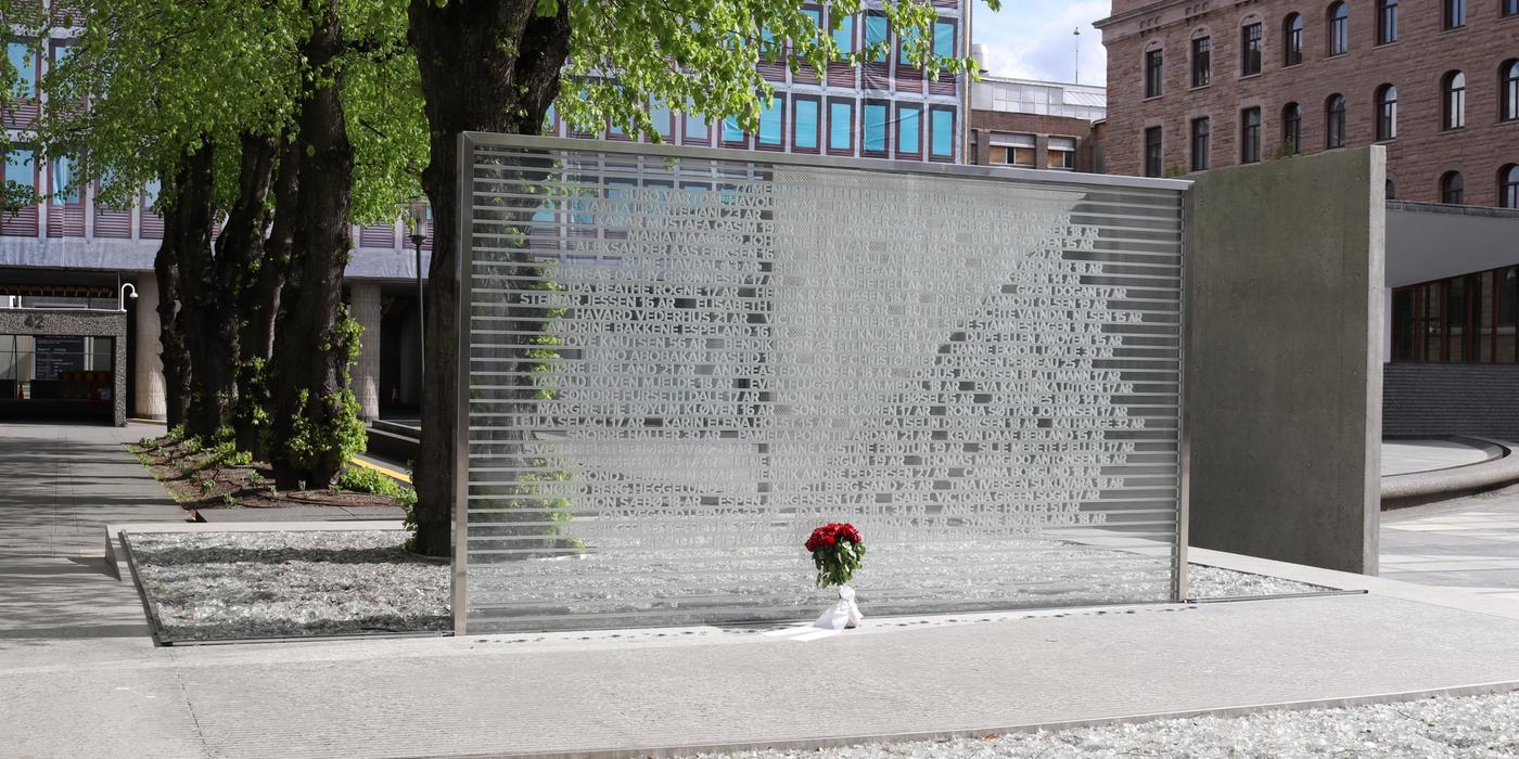 Minnesmerke formet som en glassvegg med navn på de drepte 22. juli innrisset. Veggen står foran Høyblokka, et grått betongbygg med presenning foran med bilder av vinduene på Høyblokka på, sånn at det ser ut som et vanlig bygg. Foran og bak veggen er det bed med glasskår i.