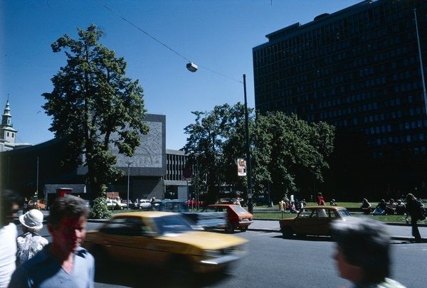 Cityscape of walking people and driving cars in front of a lawn  with seated people. In the background, a tower, a concrete buildings with engraved decoration and an office building.