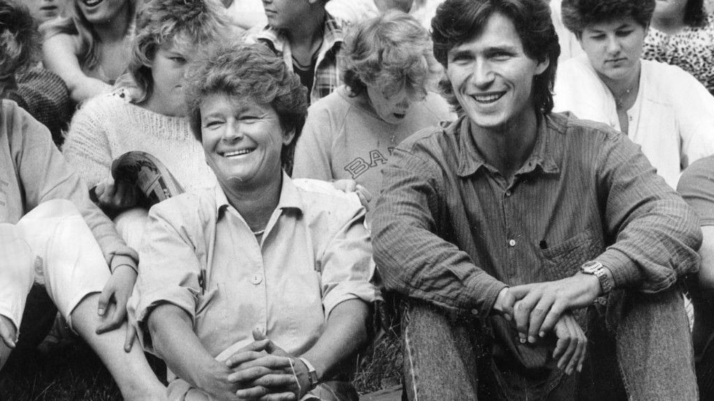 Black and white photo of smiling woman and man sitting on the ground. Several people are sitting in the background.