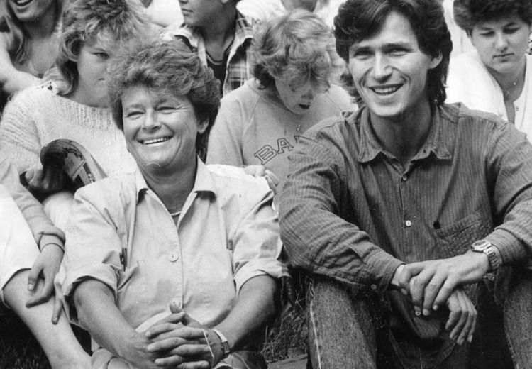 Black and white photo of smiling woman and man sitting on the ground. Several people are sitting in the background.