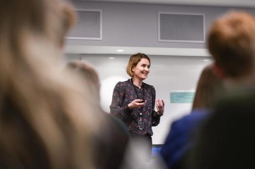 A standing woman speaks while gesticulating. She is seen through blurred shoulders from the seated audience.