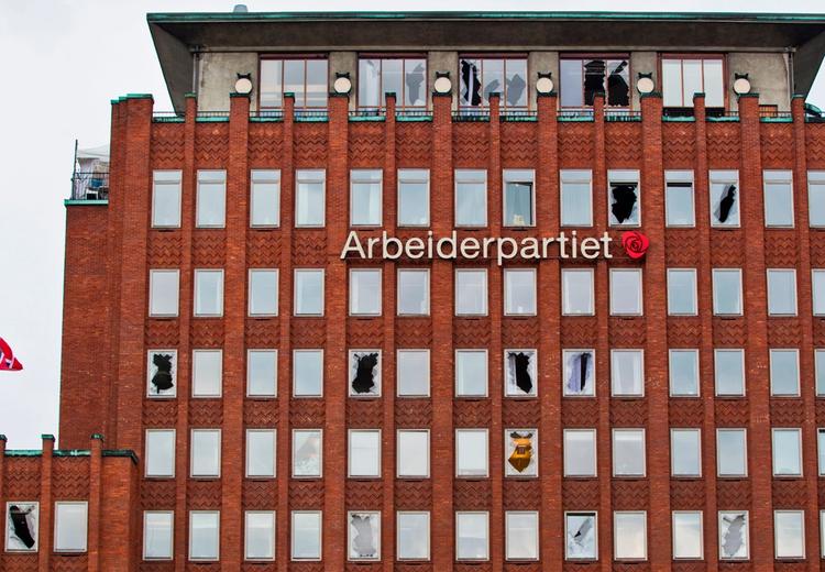 Kontorbygg med skadde vinduer, og med påskrift "Arbeiderpartiet" og deres logo. Flaggstang med det norske flagget på halv stang. 