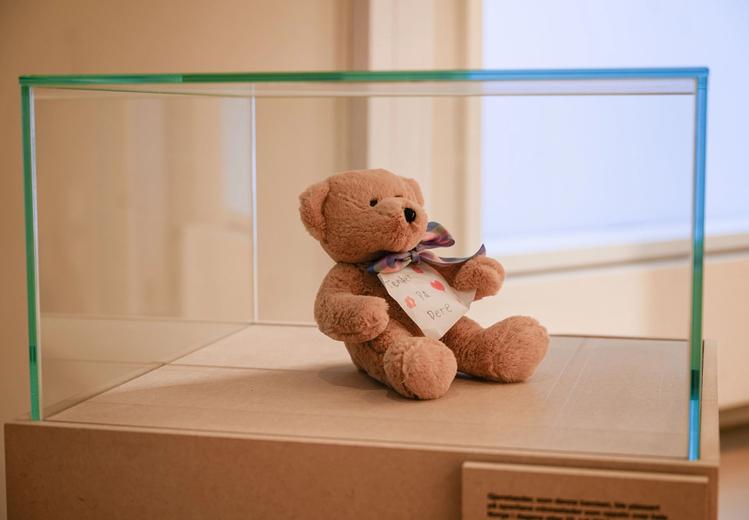 Brown teddy bear with a bow around the neck and a note with illegible writing. The teddy bear is in an exhibition stand.