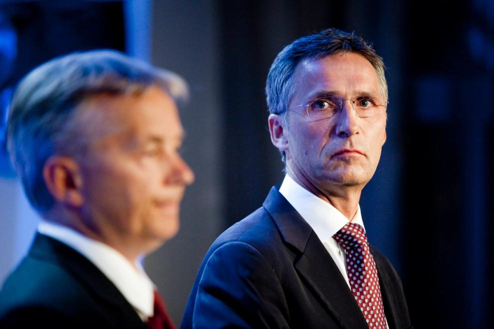 Two men side by side wearing suits and ties. The first man is out of focus, the second looks to his right with a serious face. Closeup from the chest. Dark blurred background.
