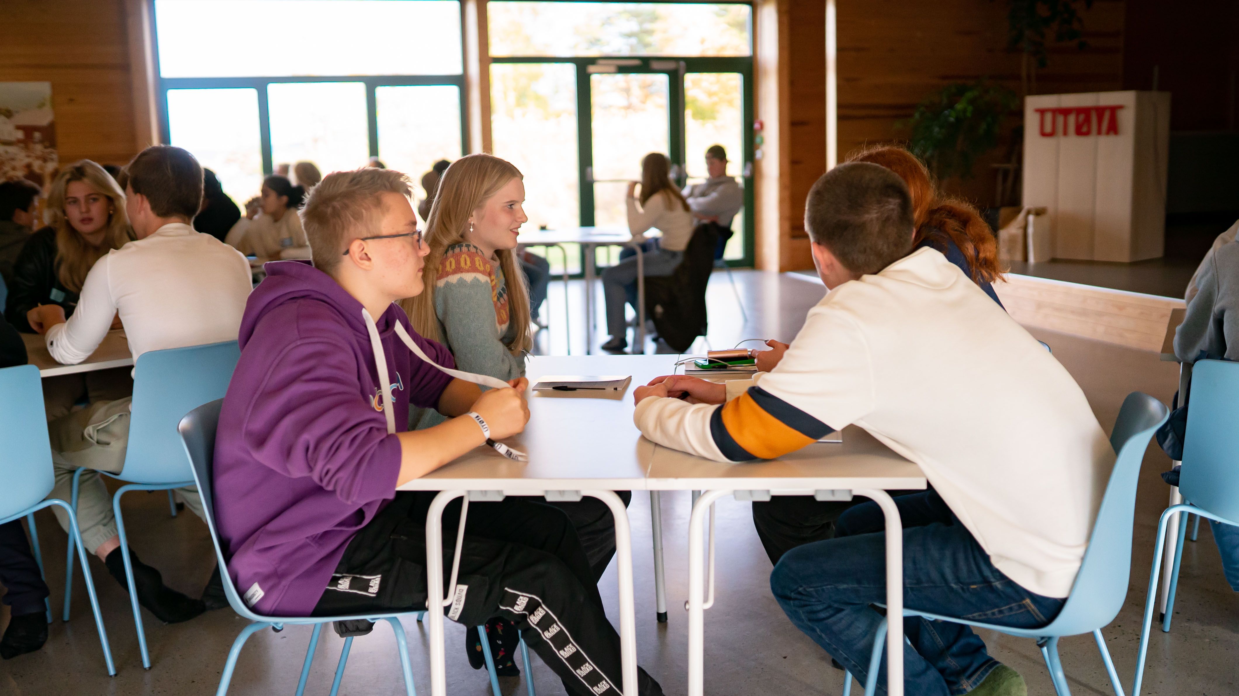 Fire ungdommer sitter rundt bord. Flere ungdommer ved bord synes i bakgrunnen. I bakgrunnen vindusrekke er stor lyskilde. Et skilt med påskrift "UTØYA".