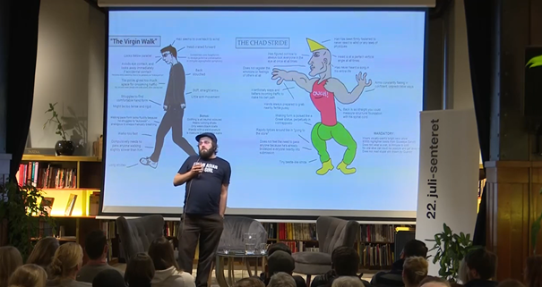 Photo of a man dressed in black standing in front of a large screen with two male figures. The audience sits and looks at the man standing. In the background a bookshelf can be seen behind the screen, to the right of the screen there is a roll-up with the 22 July Centre's logo.