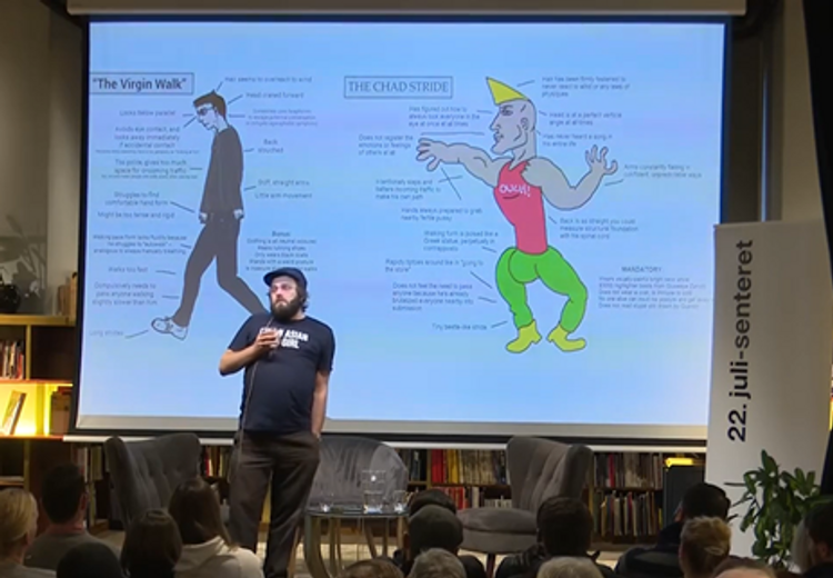 Foto av en sortkledd man som står foran en stor skjerm med to mannlige figurer. Publikum sitter og ser på mannen som står. I bakgrunnen en bokhylle ses bak skjermen, til høyre av skjermen er det en roll-up  med 22. juli-senterets logo. 