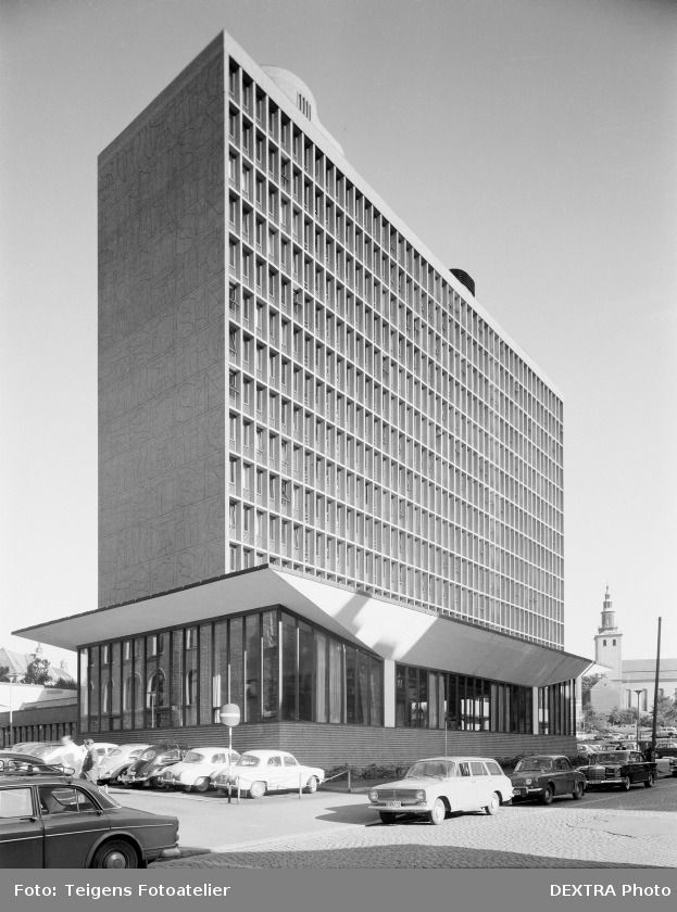 Black and white photograph of highrise building. A low rise pavillion in front. Cars parked outside.