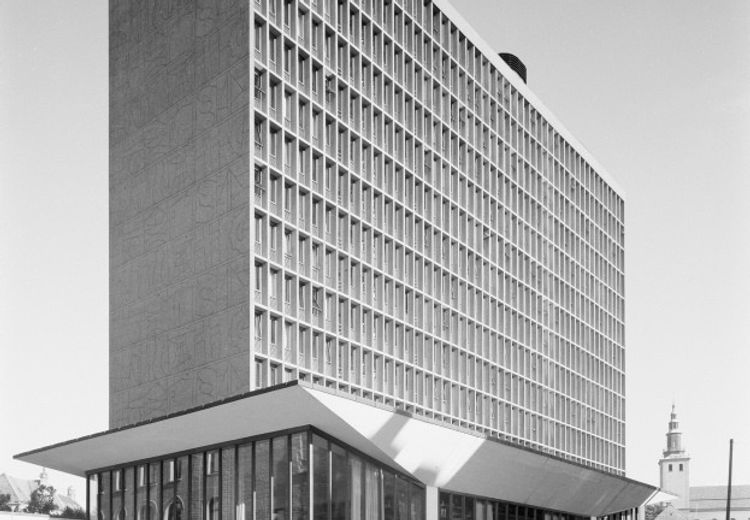 Black and white photograph of highrise building. A low rise pavillion in front. Cars parked outside.