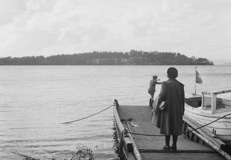 Black and white photo of a woman standing on a wooden pier with her back to the camera. In fornt of her, a boy prepares a small boat with a flag. There is an island in the backgorund.